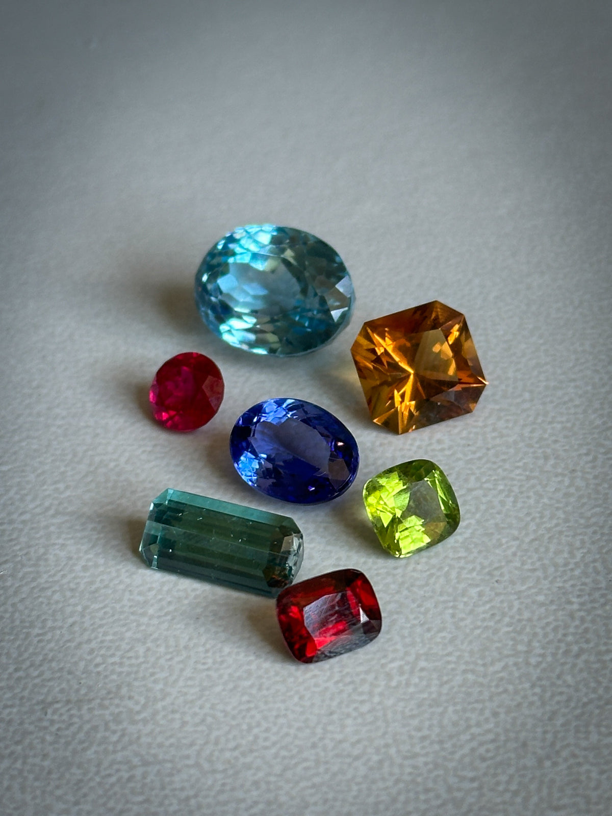 a mixture of faceted gemstones sitting on a white table - oval blue topaz, round ruby, square citrine, oval tanzanite, cushion cut peridot, baguette cut tourmaline, and cushion cut garnet